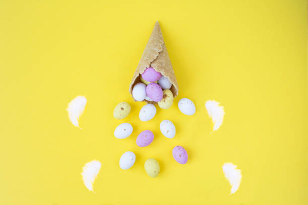 istock Ice cream cone with colored easter eggs and white feathers on the yellow background. Top view. Copy space. 1452362243