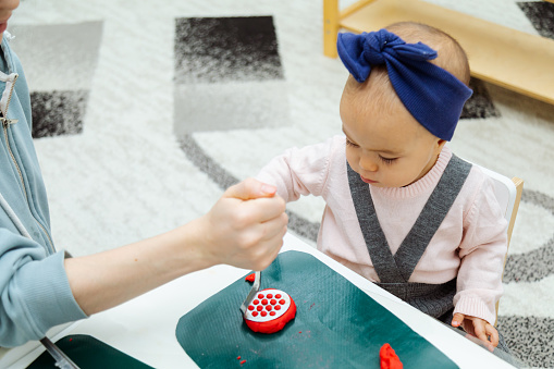 Mother teaching toddler to make shapes with colorful modeling clay or Play-Doh on a table in the kindergarten