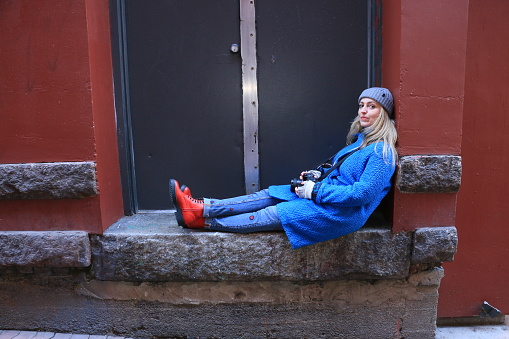 A Caucasian Polish woman resting in a doorway from taking pictures with her camera. She is wearing a gray toque, blue coat, jeans and red shoes. The building is red brick with a black door.