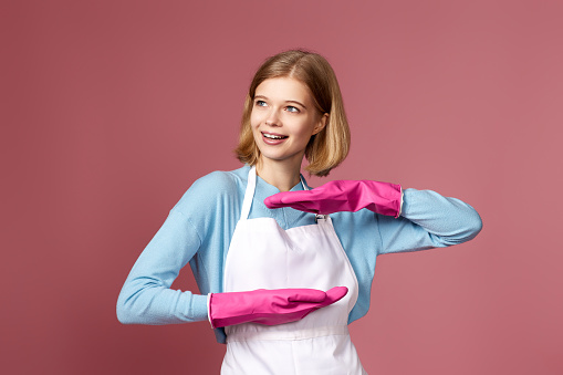 beautiful blonde woman in pink gloves and cleaner apron showing measure symbol gesture on pink background. cleaning