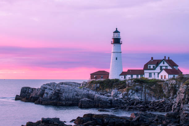 A lighthouse on a small island surrounded by calm waters with a forested coastline in the background.