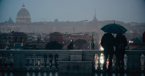 Tourists with umbrellas during a rainy day in Rome, enjoying the view from Pincio to Piazza del Popolo, with domes and cityscape.