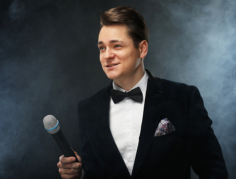 Stylish young man in a tuxedo holding a microphone, posing against a dark background with smoke, actor, singer, show, host of the event. Party concept.