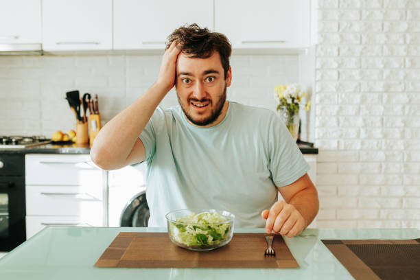 istock Front view looking at camera young guy holds head in front of him lies a fork and a plate of salad. 1452410556