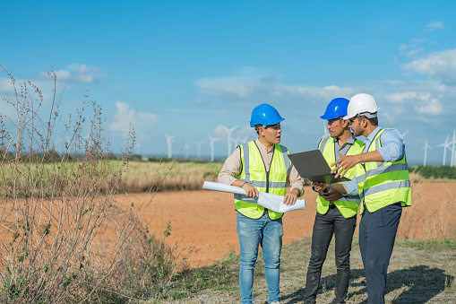 Asian engineer working in fieldwork outdoor. Workers check and inspect construction and machine around building project site. Wind turbine for electrical of clean energy and environment sustainable.