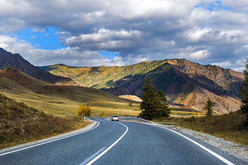 Serpentine asphalt road among high snow-capped mountain peaks, yellow desert, autumn green forest and blue sky. Car on the highway against the background of a mount landscape.