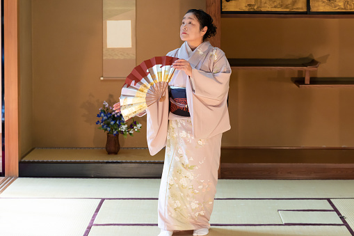 Elderly Japanese woman, with bunched hair, in flower-patterned kimono, dancing Japanese traditional dance with decorated fan in Japanese room