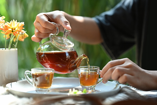 Cropped view of young man pouring traditional tea from transparent teapot into a glass cup.
