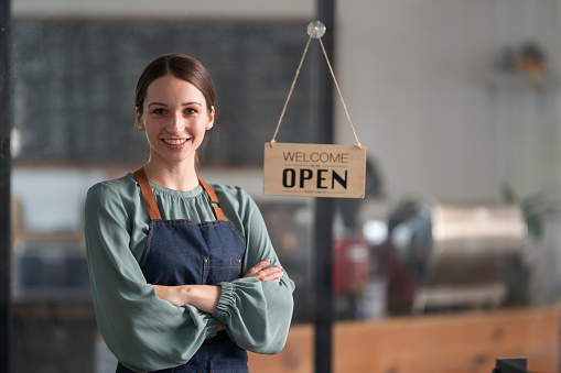 A woman who runs a restaurant and coffee shop business stands welcome confidently with her arms crossed. Hang a sign to open the restaurant. Small business concept.