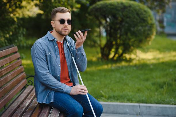 istock Young blind man with smartphone sitting on bench in park in city, calling. 1452456521