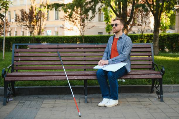 istock Visually impaired man with walking stick, sitting on bench in city park. Copy space 1452457412
