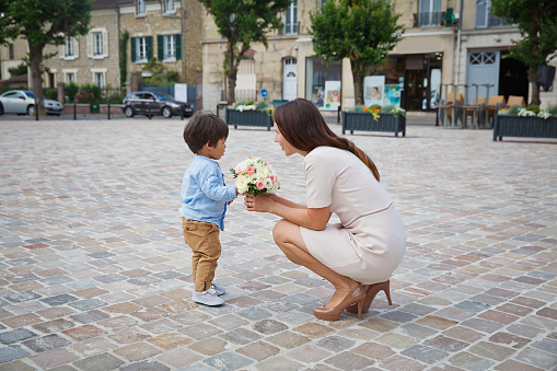 Mixed race boy little son congratulating his mom and giving her flower bouquet, they hugging and laughing together. Family holidays concept