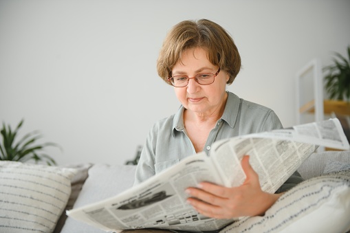 Senior lady reading her newspaper at home relaxing on a couch and peering over the top at the viewer.