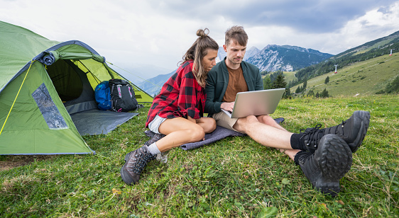 Photo of young couple using laptop while camping in mountains. Modern lifestyle and nature concept.
