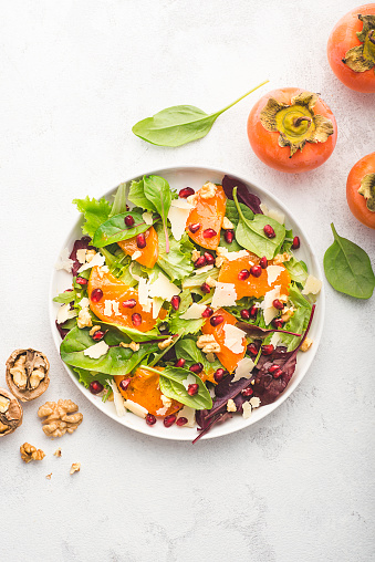 Juicy unusual salad with persimmon, pomegranate, fresh spinach, various leaves, pecorino cheese and walnuts. Sweet and salty tastes, winter and autumn salads, gluten free recipe. Top view