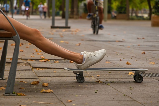 A girl sitting on a bench in the pedestrian street and putting her feet on the skateboard