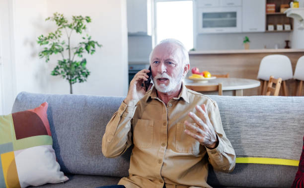 istock Grey-haired senior hipster man in casual shirt using smartphone sitting on the sofa at home. Elderly guy enjoys online chatting, spends time in social networks, using phone and smiles. 1452485196