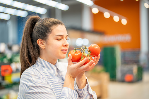 Young woman holding and smelling fresh organic tomatoes with the eyes closed at the vegetable section in a supermarket. Making healthy nutritional choice. Woman enjoying the smell of organic raw vegetables during grocery shopping.