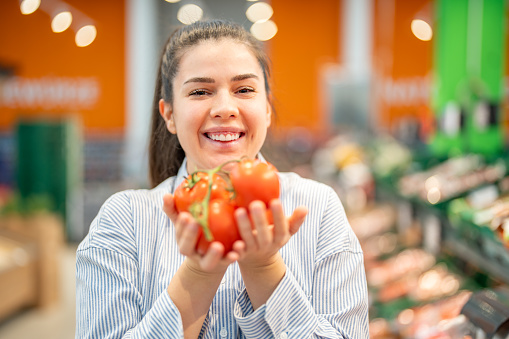 Smiling young woman holding fresh tomatoes making healthy choices during grocery shopping in a supermarket. Concept of a healthy diet.