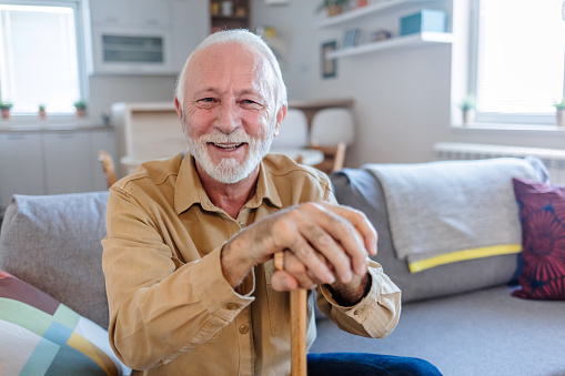 Shot of an elderly man sitting on the couch with a walking stick at home. Senior man leaning in his walking stick at home.