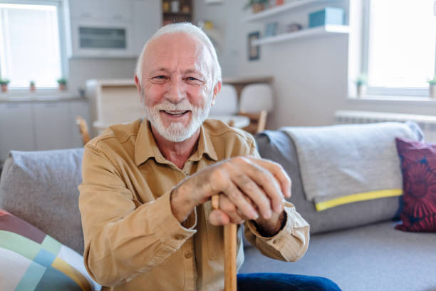foto de um homem idoso sentado no sofá com uma bengala em casa. homem sênior inclinado em sua bengala em casa. - 70 anos - fotografias e filmes do acervo