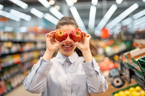 Young woman having fun while doing grocery shopping in the supermarket, standing towards the camera, holding two apples and smiling. Healthy eating concept.