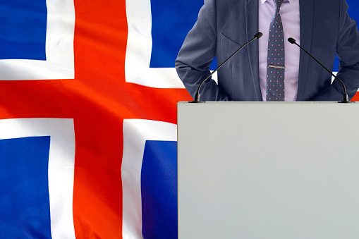 Tribune with microphone and man in suit on Iceland flag background. Businessman and tribune on Iceland flag background. Politician at the podium with microphones background Iceland flag. Conference