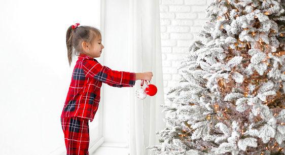 A girl in checkered pajamas holds a Christmas ball in her hand while decorating an artificial Xmas tree. The child is preparing for the new year