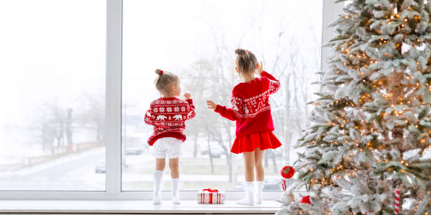 Two little girls in red patterned sweaters are standing on the windowsill by large window with Christmas tree watching falling snow outside. Children waiting for  new year and Xmas miracles