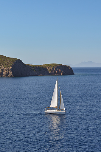 Sailing yacht with white sail on a calm sea with coastline in the background