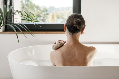 Concept of spa procedure at home. Beautiful woman looking at window while she taking a bath. Girl resting in bathroom, sit in hot water. Back rear view of female washing body with scrubber or sponge