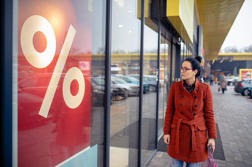 Woman walking outdoors in a city. She is window shopping and looking at a store sale advertising or marketing apparel and clothing on a display glass.