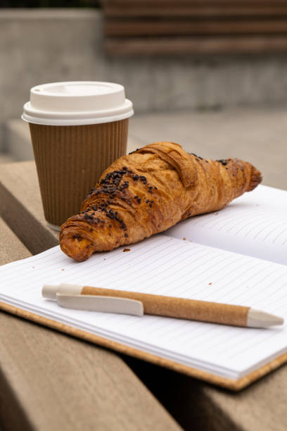 istock Fresh baked chocolate croissant on paper cup with lid for tea to go. Empty paper notebook. Freelancer's place of work. Study and work online. Remote business education. 1452499352