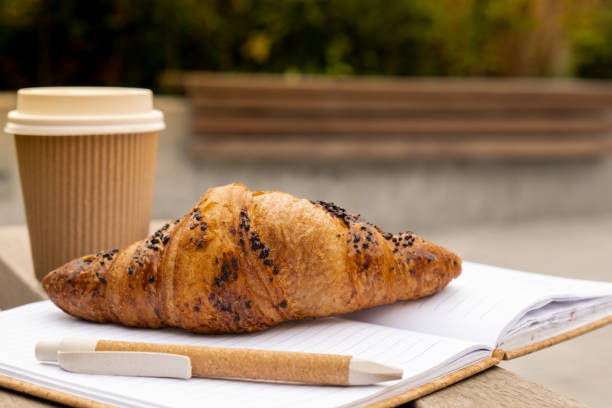 istock Fresh baked chocolate croissant on paper cup with lid for tea to go. Empty paper notebook. Freelancer's place of work. Study and work online. Remote business education. 1452499456