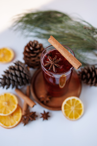 Istanbul, Turkey-December 22, 2022: Red colored, clove grained, star anise winter tea in a tall glass decorated with cinnamon sticks on a light gray background. The tea is surrounded by a coniferous pine tree branch, cones, cinnamon sticks and dried orange slices. Shot with Canon EOS R5.