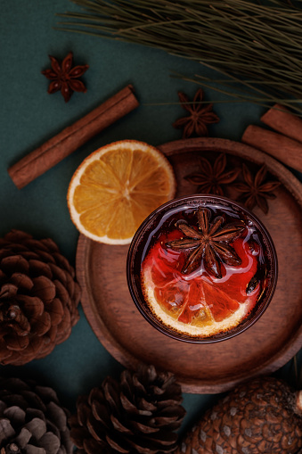 Istanbul, Turkey-December 22, 2022: Red colored, clove grained, star anise winter tea in a tall glass decorated with dried orange slice on a teal background. The tea is surrounded by a coniferous pine tree branch, cones, cinnamon sticks and dried orange slices. Shot with Canon EOS R5.