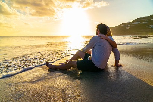 A beautiful young couple sits on a tropical beach and enjoys the view to the turquoise ocean during the sunset