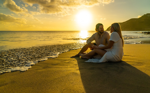 A beautiful young couple sits on a tropical beach and enjoys the view to the turquoise ocean during the sunset