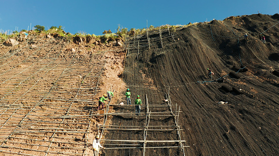 Protection of road from mountain slough, rockfall with metal accumulative restraining net fences. Workers constructing anti-landslide concrete wall prevent protect against rock slides. Rockfall protection. Workers strengthen the slope of the mountain with steel grid preventing rockfall and landslide on the road.