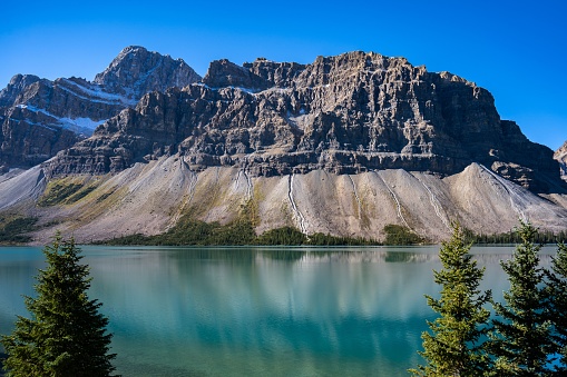 A magnificent view of a transparent mountain lake's surface, with an evergreen forest and a rocky mountain in the background, on a sunny day