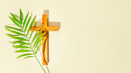 Palm Sunday Holiday. Wooden cross with palm leaf on light background with copy space. Religion background suitable for faith religion, christian holidays, Easter, Redeemer, the Feast of Corpus Christi