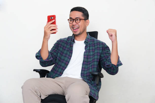 Asian man sitting in an office chair showing excited when looking to his phone Asian man sitting in an office chair showing excited when looking to his phone kantor stock pictures, royalty-free photos & images