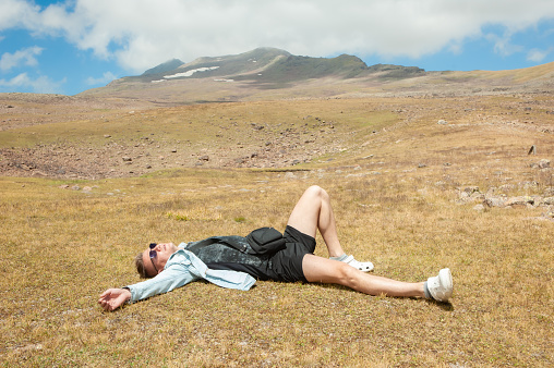 A young man in a denim jacket and shorts lay down on a lawn in the mountains and spread his arms to the sides. A man escaped on vacation and is happy to be in nature in the mountains enjoying life