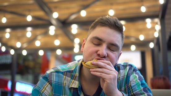 Hungry man is actively eating a burger. A young man in a cafe eats a burger and drinks coffee. Close-up of a man's face. 4k