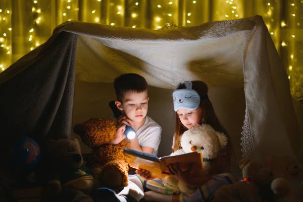 istock Two children with flashlight read a book under a blanket as a tent. 1452531684