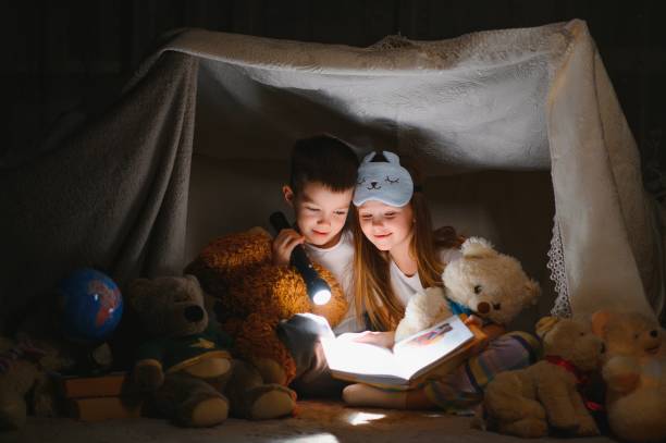istock Two children with flashlight read a book under a blanket as a tent. 1452531693