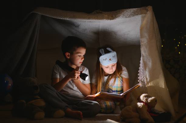 istock Two children with flashlight read a book under a blanket as a tent. 1452531737