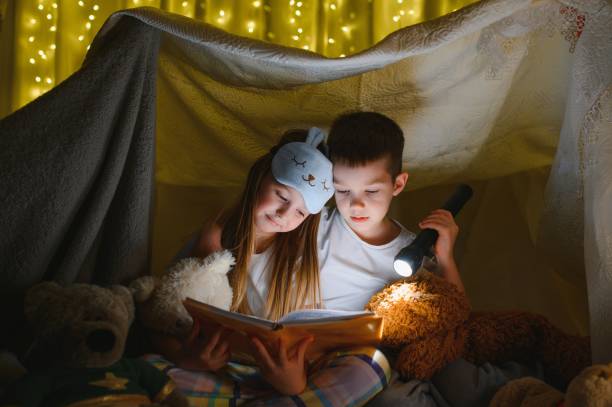 istock Two children with flashlight read a book under a blanket as a tent. 1452531884