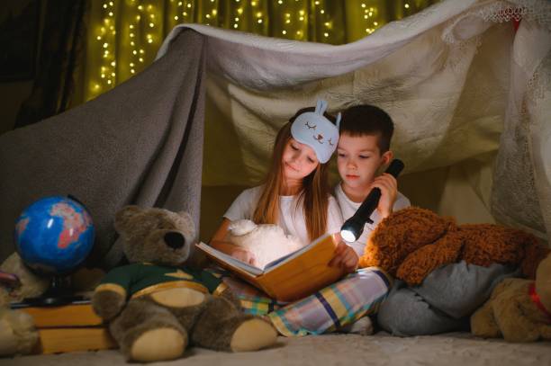 istock Two children with flashlight read a book under a blanket as a tent. 1452531922