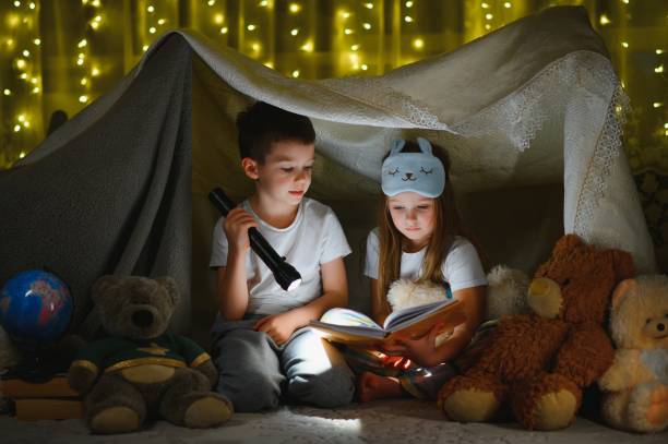 istock Two children with flashlight read a book under a blanket as a tent. 1452532067